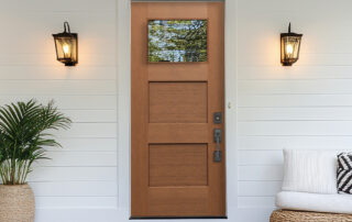 Modern wood front door with small glass window, black hardware, and porch sconces on a white exterior wall.